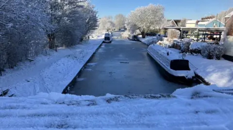 A frozen canal, with snow on top of boats and a bridge