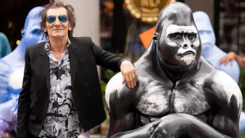 PA Media Image of a Ronnie Wood in a patterned shirt and black jacket and wearing sunglasses standing next to a statue of a gorilla.