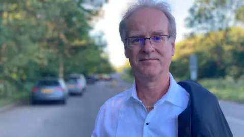 Simon Bentley stands on a rural road wearing a dress shirt and blazer on one shoulder.