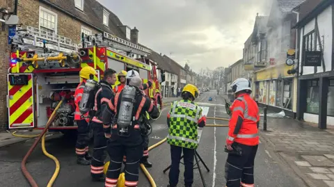 The picture is looking down a street that has buildings either side. Parked up on the left is a fire engine, which is red and yellow, with hoses coming out of the back. There are seven members of the fire service standing near the fire which is coming out of the building on the right. 