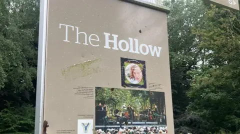 A tall beige upright sign which has the words 'The Hollow' at the top. There are small images of Van Morrison, one from the cover of his album Astral Weeks, and the other taken from a live performance. Behind the sign are lush green trees.