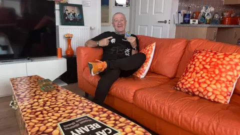 Captain Beany at home with his baked-bean decorated coffin that he uses as a coffee table