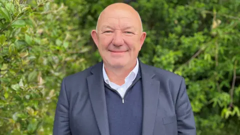Head and shoulder shot of a smiling Andy Sloan, who is bald, smiling at the camera. He is wearing a navy suit jacket, navy half-zip jumper and a white shirt with the top button open. Green shrubbery is visible in the background.