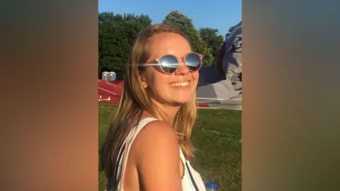 Julie Devey A young girl with blonde hair who appears to be standing in a field. She is looking at the camera and smiling, wearing a white vest and sunglasses.