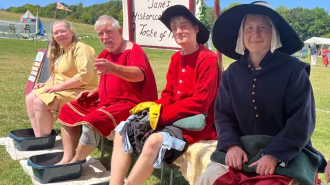 Four historical re-enactors sitting on a bench in the sunshine, they all have their feed dipped in washing up bowls filled with water. The sign, Jane' Historical Kitchen can be seen in the background.