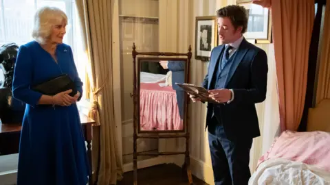 Charles Dickens Museum Ollie Dickens, great-great-great grandson of Charles Dickens, with HM The Queen at the Museum. They are standing in a bedroom, by a mirror. Ollie is holding a clipboard, whilst the Queen holds a handbag and watches Ollie reading