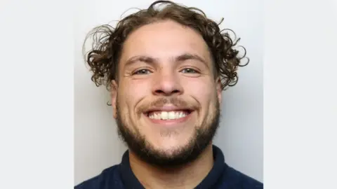 Wiltshire Police A police mugshot of Reuben Brown, wearing a dark blue polo shirt and standing against a grey background. He has curly brown hair and a beard, and is grinning at the camera.