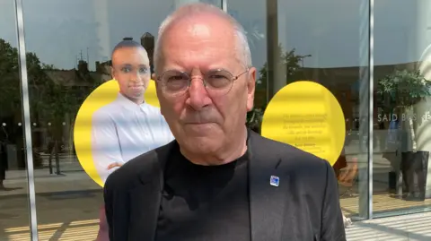 Lord Hendy wearing a dark blazer and black t-shirt whilst standing in Oxford. He is bald and wears thin-wire glasses.