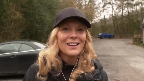 Lisa Lewis smiling looking at the camera. She is wearing a cap and earphones. She has curly blonde hair. It looks like she is out walking. Behind Lisa is a carpark with two cars parked. 