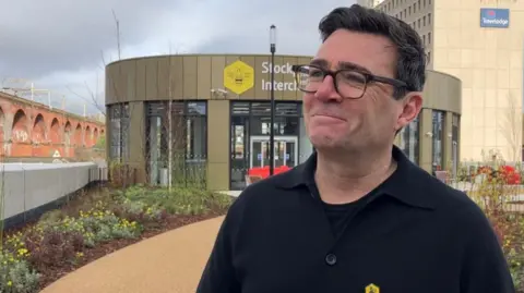 LDRS Andy Burnham smiles at the camera outside Stockport Interchange. He has dark short hair and is wearing dark rimmed glasses and a dark shirt and t-shirt. Railway arches and a Travelodge are behind him.