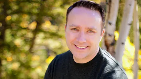 Greg Daily Greg Daily, wearing a black t-shirt, smiles at the camera, in front of trees