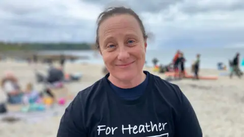A woman with brown hair tied back. She is wearing a black top that says 'For Heather'. Behind her the background is blurred but there is a sandy beach with people walking and the sea. 