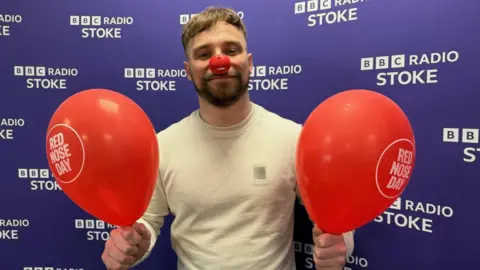 A man with short hair, a beard and a white top, smiles as he wears a Red Nose Day red nose and holds two red balloons in the BBC Radio Stoke studio.