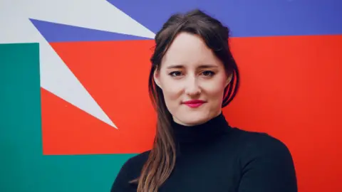 Mathlide Davis Dr Mathilde Pavis is looking directly into the camera. She has long brown which is draping over her right shoulder. She is standing infront of a multicoloured wall. 
