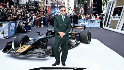 Getty Images Brad Pitt standing in front of a black and gold F1 car at a movie premiere.
