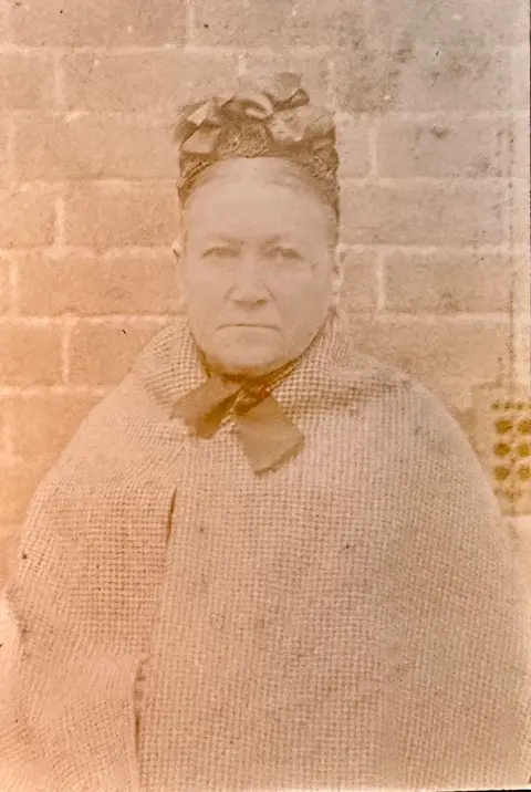Thames Valley Police Museum A sepia close-up photograph of a stern faced heavily built woman in her fifties with her hair parted in the middle and mostly hidden by a hat made out of lace with bows on the top. She is wearing a checked shawl and standing in front of a brick wall.