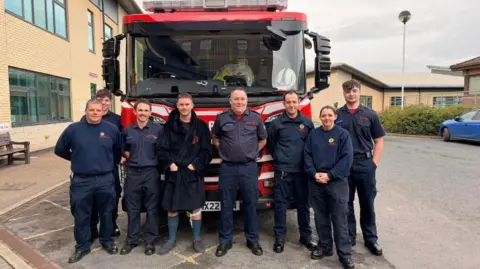 Shropshire Fire and Rescue Service Eight people are standing in front of a red fire engine. A building is on the left of the photo and a smaller building is in the background on the right.