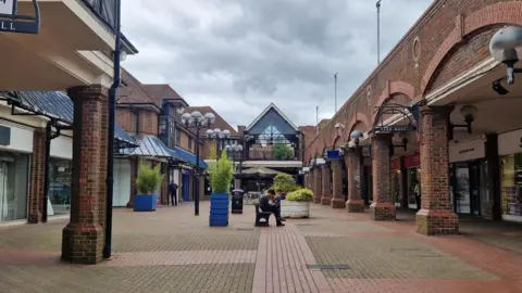 An outdoor colonade style shopping centre. There are several shops in the picture as well as large pot plants and a bench with a young man sitting and reading. The sky is overcast.