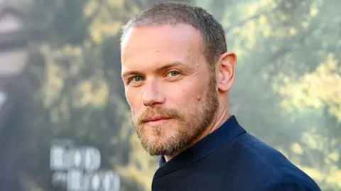 Getty Images Sam Heughan looking straight at the camera while standing in front of a blurred backdrop.