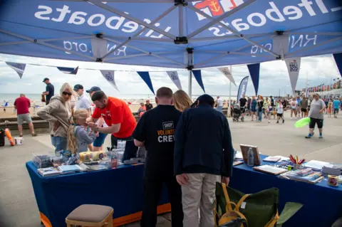 RNLI / Mike Milner A blue RNLI marquee with three people standing near chairs with someone's shirt saying "shop volunteer crew". There are leaflets on the tables and a man in an orange top is putting something around a blonde-haired girl's neck on the other side of the stall. You can see lots of people walking along the pier.
