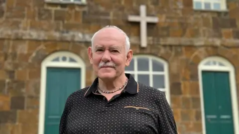 Carol Weston/BBC man looks at the camera. He wears a short -sleeved black polo shirt. He stands in front of a two -storey stone building with green doors and crosses the wall. 