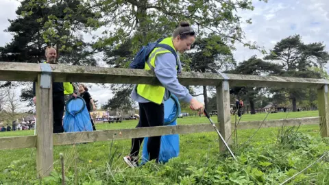 A man and a woman in yellow hi-vis vests are collecting rubbish with litter pickers and putting it in blue bags. They are working at the edge of a park with large trees behind them.