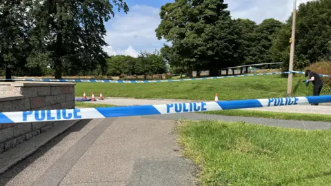 A strip of blue and white police tape across the image. There is a grey wall on the left hand side and a grey path going through the middle. A patch of green grass is on the right hand side. On the far right of the image, a police worker with long, dark hair and wearing dark clothing and blue protective gloves is partially bent over and is holding something in their hands.