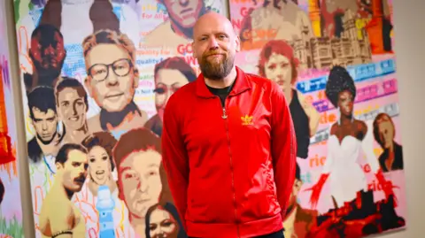 Out North East Spray paint artist Chris Fleming, also known as ID4A, stands in front of his artwork. The art is made up of brightly coloured panels with figures from popular culture and landmarks from around the North East. Fleming wears a red sports jacket.