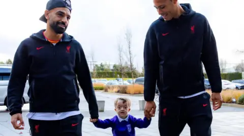 Family photo Mo Salah, with a trimmed black beard and who is wearing a baseball cap with the peak turned backwards and black zip-up Liverpool FC branded jacket and jogging bottoms, holds the left hand of a little boy while Virgil Van Dijk, who is taller and wearing the same tracksuits, holds his right hand. 