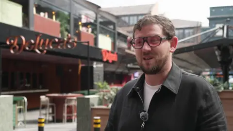 A man in his thirties, with light brown hair, a brown beard and wearing a black half zip jumper and tinted glasses, speaks to the camera in front of an outside seating area in front of a bar. 