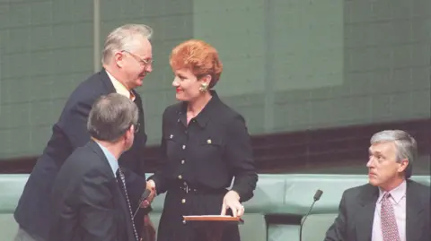 Getty Images A woman with short red hair wearing a black dress shakes hands with a man wearing a suit as two other men in suits look on 