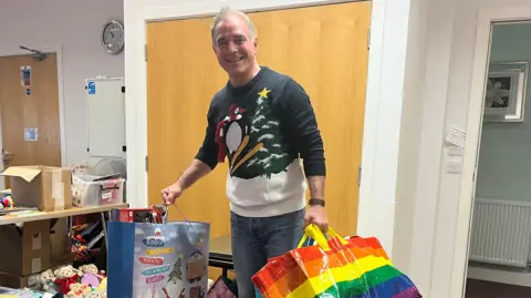 ELLEN KNIGHT/BBC Mike pictured looking into the camera and smiling. He's stood by the door holding three large gift bags full of food and toys. He's wearing a knitted Christmas jumper that depicts a skiing penguin and a Christmas tree, and blue jeans. 
