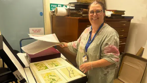 Gilly Hope/BBC Gilly Hope, a middle-aged woman with glasses and tied-back brown hair, wearing a shirt with large, pink flowers on it. She is stood looking through a collection of watercolour paintings of flowers by Sunderland artist Charles Muir.