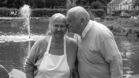 Martina Penna A black and white photograph of a white haired lady in an apron, smiling at the camera. A balding man in a v neck sweater and shirt is kissing her on the cheek. They are both by a pond in a park