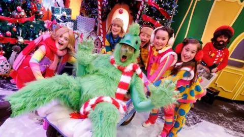 Andres Poveda A man dressed in a woolly green costume and green face paint sits in a blue basket on wheels. He wears a red and white striped scarf and green helmet. Six kids dressed in Christmas costumes stand around and behind him. There is a large green Christmas tree to the left of the frame. A green and gold miniature house is behind them to the right of  the frame 