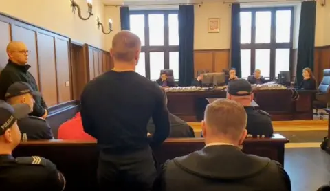 View of the courtroom wit the defendants seen from behind, police on either side 