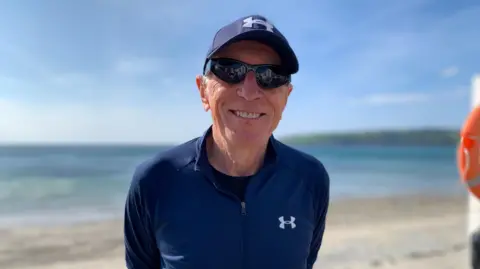 Mark is wearing a navy quarter-zip top with a navy cap and sunglasses. He is smiling in front of a calm Laxey beach on a sunny day.