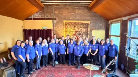 Sam Scherdel Sam Scherdel pictured with members of Hull NHS Choir inside a recording studio. Members of the choir are wearing matching blue tops and there is a drum kit and keyboard in the foreground.