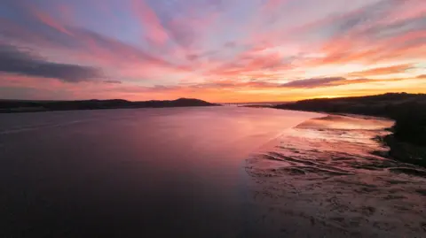 Frazer Grant A wide river or estuary at sunset, with smooth water reflecting pink and orange clouds and a sandy shoreline curving along the right side.