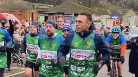 Kevin Sinfield with runners and people on bikes. He's wearing a green vest with sponsorship and a big number 7. It looks very cold.