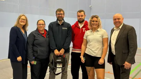 Wiltshire Council Six people standing in an indoor sports setting smiling at the camera. One man holds a defibrillator.