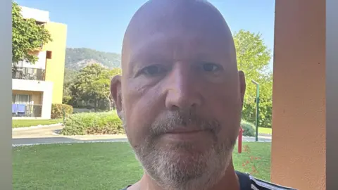 Gavin Sparks takes a selfie at his hotel. He has a bald head and a short grey beard. He is wearing a black Adidas t-shirt with white stripes on his arm.