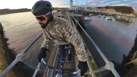 Mike Humphreys Mike Humphreys riding the Chopper along a narrow railed bridge over the water. There are boats, houses and trees in the background.