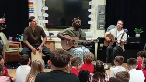 Contributed Smith sits in a chair at the front of a classroom playing a guitar. Another man sits to his right playing a guitar and a man to his left who provides percussion on a box. 