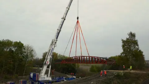 Chesterfield Canal Trust A photograph of the bridge being lowered into place by a large white crane. There are engineers helped the bridge into place. The bridge is surrounded by trees.