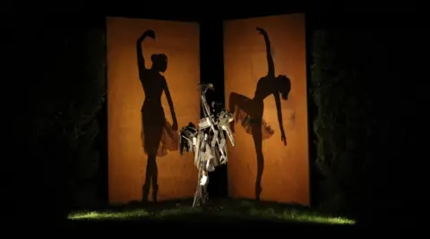 Will Budgett A metal sculpture positioned in between two large orange canvases, casting a silhouetted shadow of dancing ballerinas in the dark.