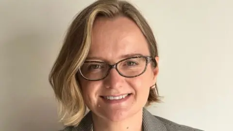 A woman with short blonde hair and glasses smiles at the camera. She is standing in front of a white wall. 