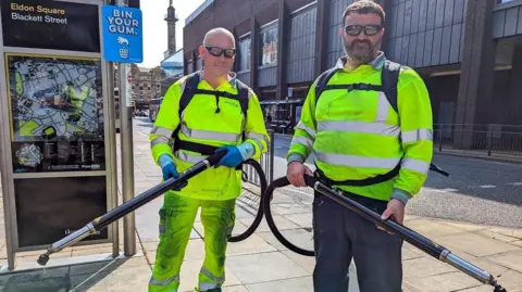 Newcastle City Council Two men wearing hi-vis clothing and gum removal backpacks. They are holding long poles attached to the backpacks and are standing on a "Bin Your Gum" sign on Newcastle's Blackett Street.
