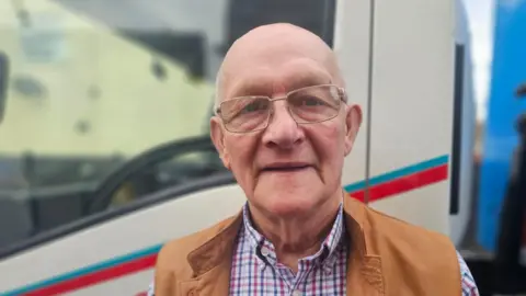 Ben Woolley smiles at the camera, he is bald and is wearing glasses. He has a checked shirt and a brown leather gilet on. 