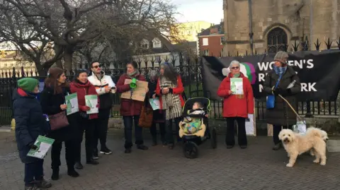 A group of people are standing outside a church in Norwich. There are 9 people, a baby in a pushchair, and a dog. They are holding signs. 